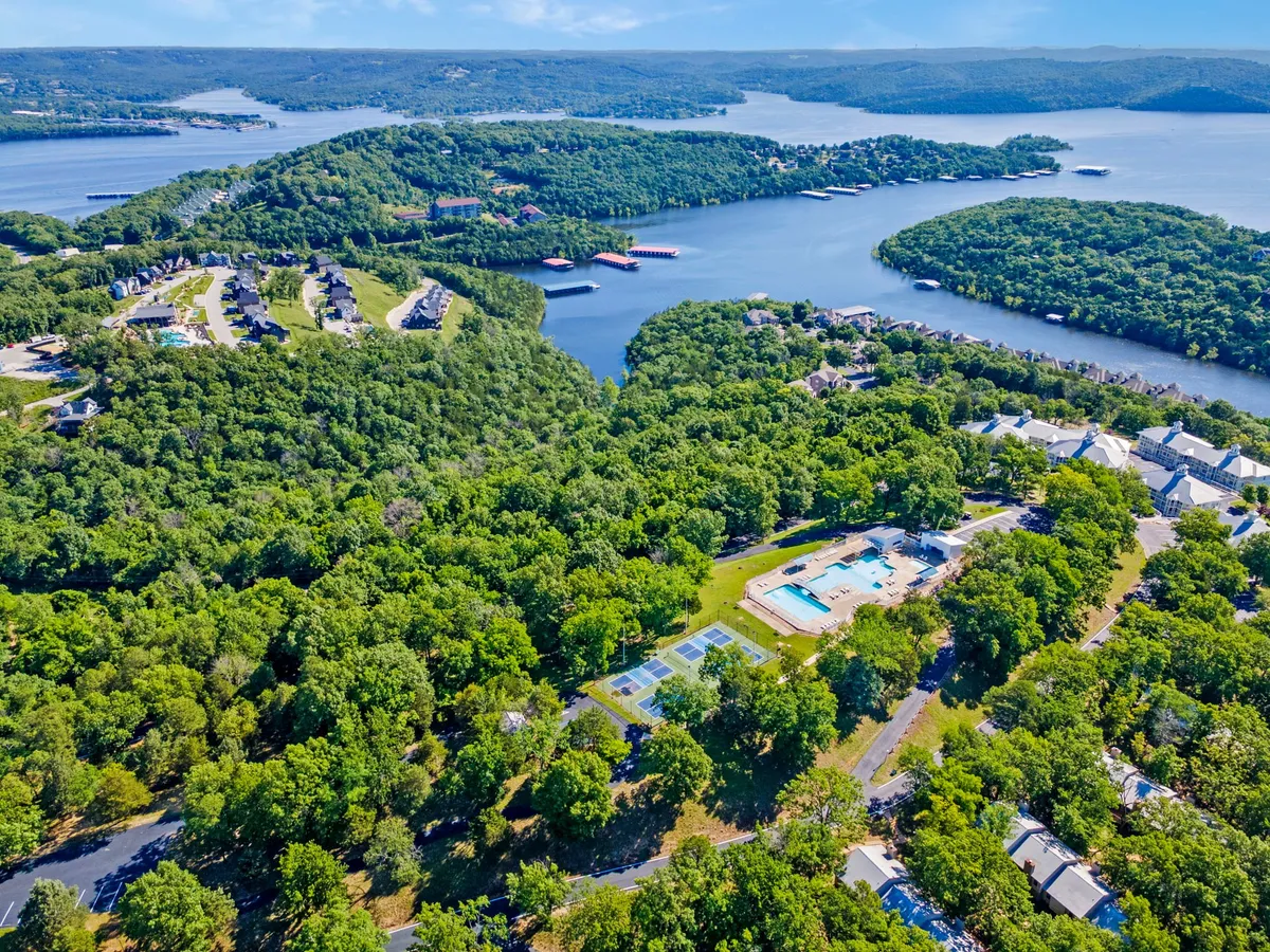 An elevated view of the expansive lake surrounded by greenery, with the swimming pool area and tennis courts clearly visible. Residential buildings are also shown, giving context to the group's home base for exploration and relaxation.