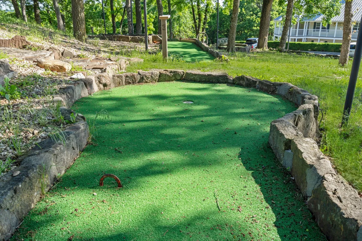 A playful exterior shot of the mini-golf course, bordered by stonework and shaded by mature trees. The course looks inviting and is clearly designed for families or groups to enjoy friendly competition in a natural setting.