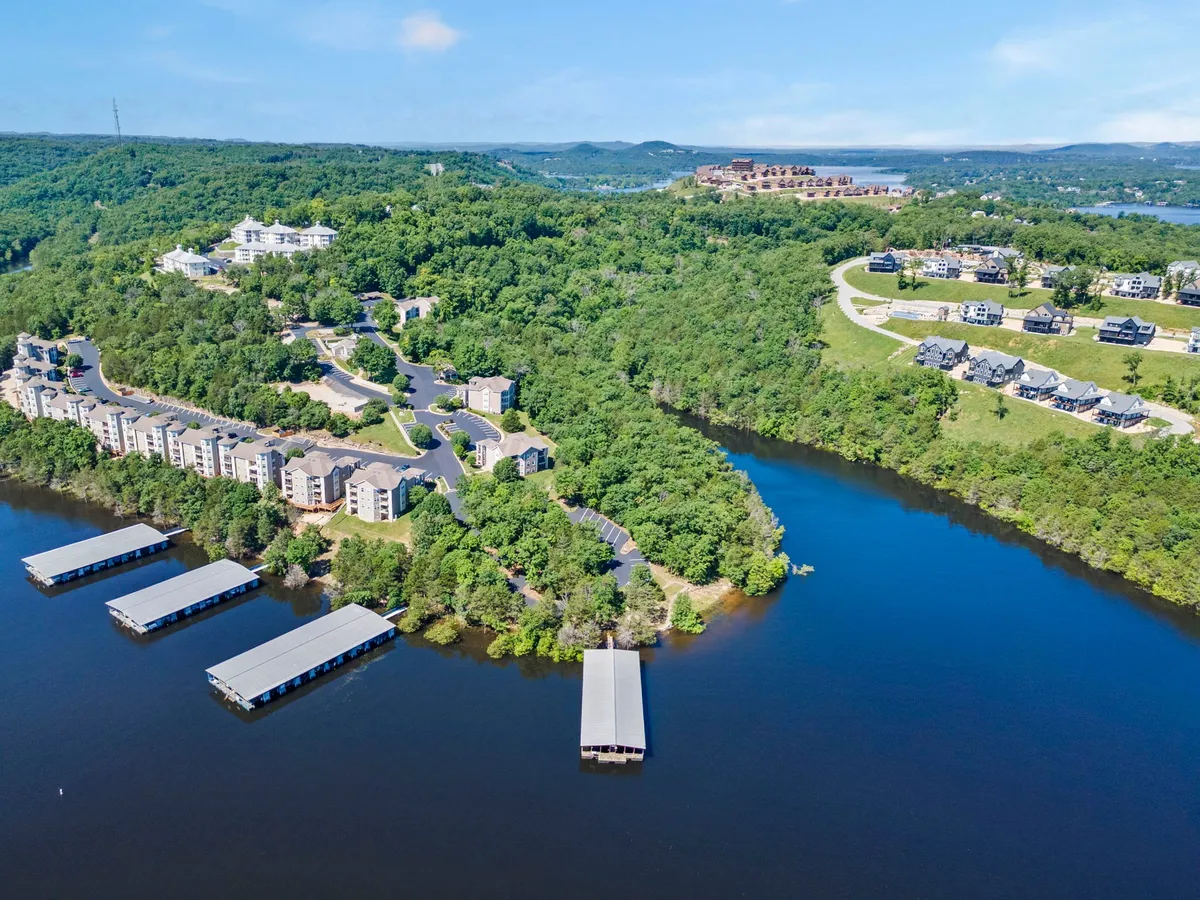 A serene view over tranquil Table Rock Lake, with luxurious waterfront properties, green hills, calm water, and visible boathouses. The image encapsulates the lakefront access, opportunities for fishing, kayaking, and boating, and the inviting natural environment.