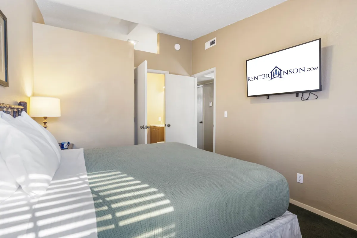 A cozy bedroom featuring a luxurious king-sized bed dressed in crisp white linens. A flat-screen TV is mounted across from the bed, and soft lighting from a bedside lamp creates a relaxing ambiance. The room has ample space and includes stylish décor touches ideal for a restful retreat.