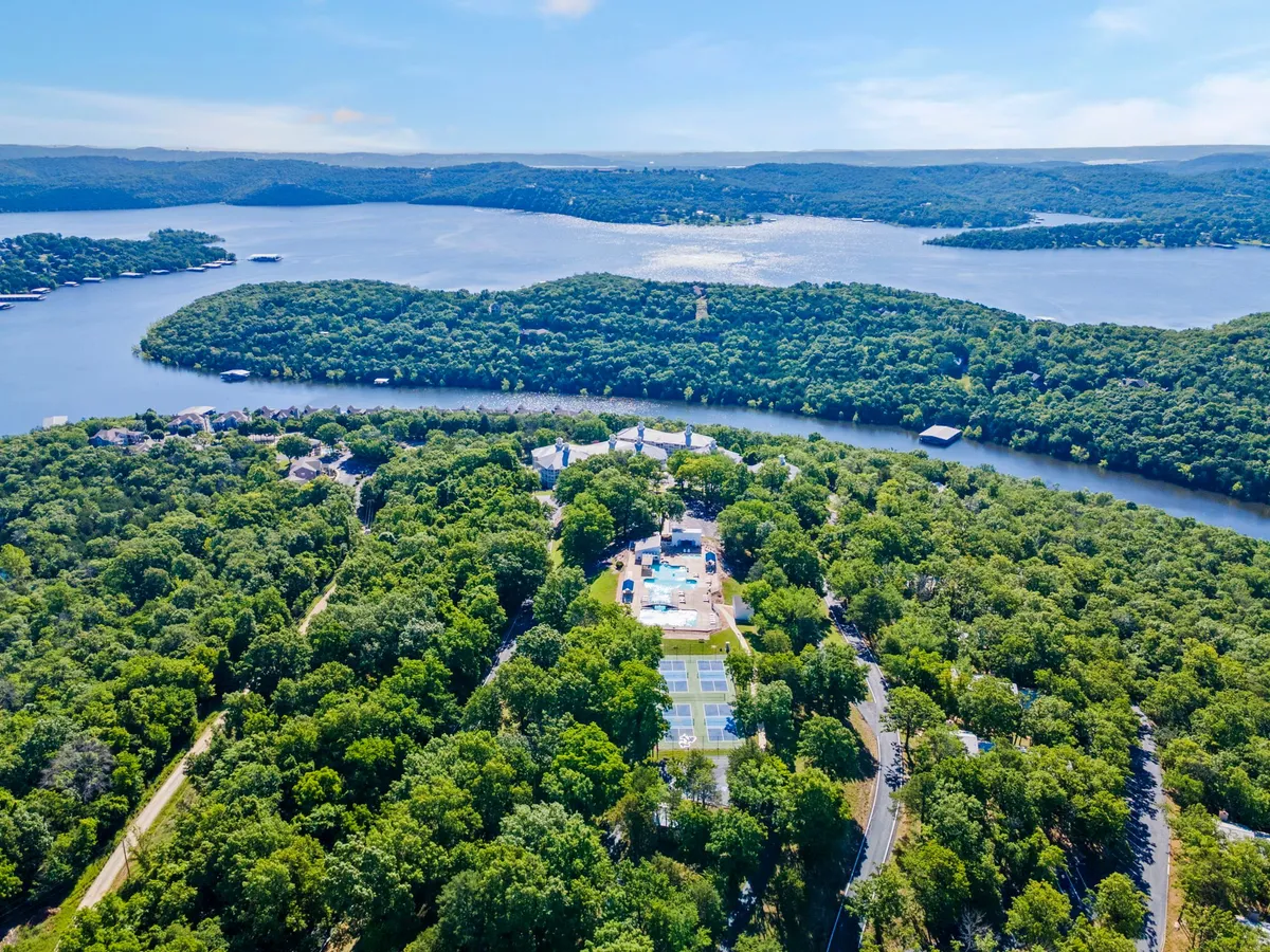 This exterior image captures an expansive water view, lush greenery, a swimming pool, and visible tennis courts. It shows the breadth of resort amenities in a scenic lakeside setting and hints at opportunities for swimming, boating, hiking, and outdoor games.