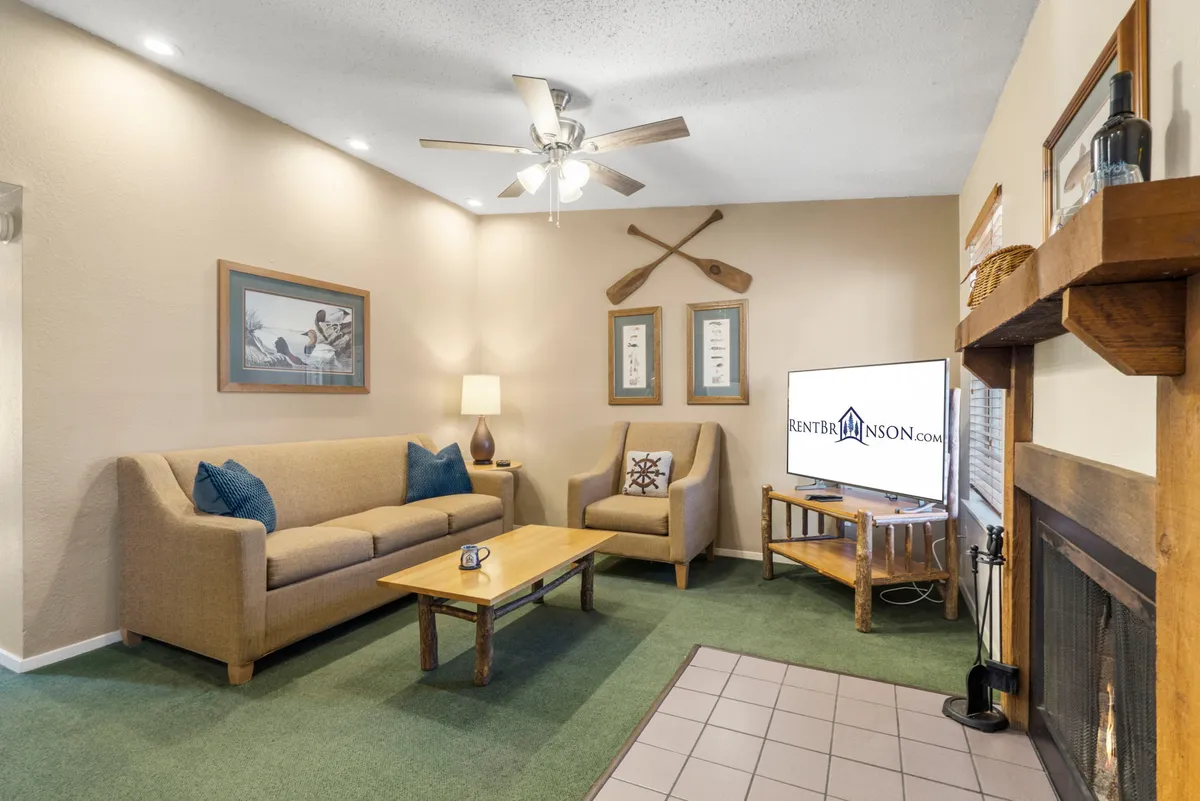 A cozy living room with a plush sofa, coffee table, a prominent fireplace, flat-screen TV, and decorative oars on the wall. The space feels inviting and is bathed in natural light, highlighting the central gathering area for relaxation or entertainment.